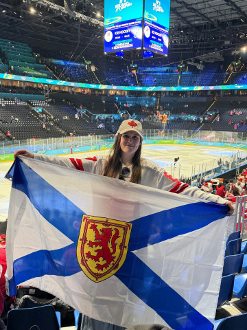 A photo of a woman with long dark hair. She is wearing a Canadian hat and jersey holding a Nova Scotia flag