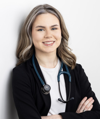 Photo of a woman with blonde curly hair. She is wearing a black jacket over a white shirt and has a stethoscope around her neck